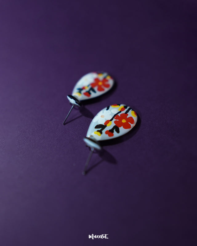 A shot of the floral earrings kept against purple background, showcasing their delicate flower design and soft pastel tones, capturing the charm of artisanal craftsmanship and subtle sophistication — Muselot.
