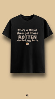 Black graphic t-shirt showcasing the statement “She’s a 10 but she’s got those ROTTEN deviled egg farts” in bold beige typography with a small egg illustration, highlighting an sarcastic and attention-grabbing humor vibe against a clean beige background – Muselot