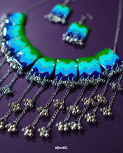Waterfall boho jewelry set placed on a deep purple background, highlighting its vibrant enamel panels in green, aqua, and navy with a touch of glitter. The silver chains and bead clusters showcase a flowing, layered design that gives a rhythmic and stylish look — Muselot.