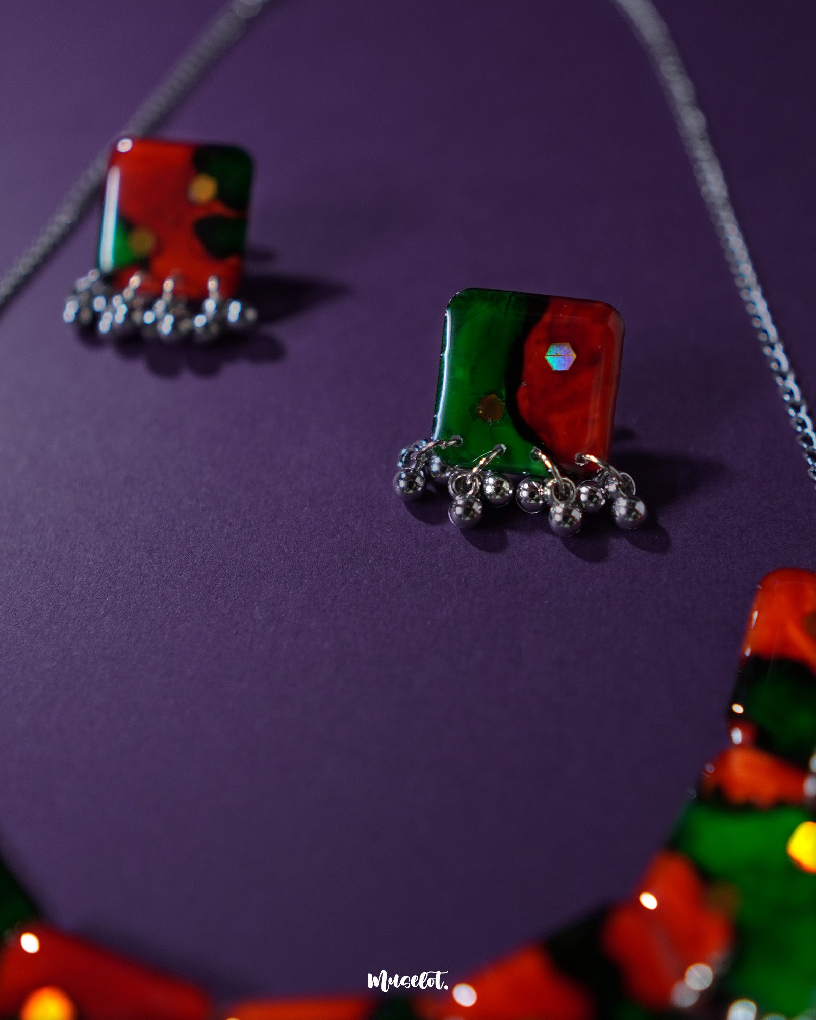 A close-up of the Surkhbaagh handmade earrings placed against a purple background, highlighting their vibrant red and green block design, glinting mirror accents, and dangling silver-toned beads that add a festive, artisanal charm — Muselot.