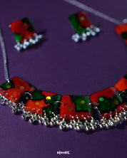 The Surkhbaagh handmade jewelry set laid against a muted purple backdrop — showcasing oxidized silver-tone earrings and a matching necklace with deep red beads, mirror detailing, and dainty ghungroos for a festive ethnic charm — Muselot.