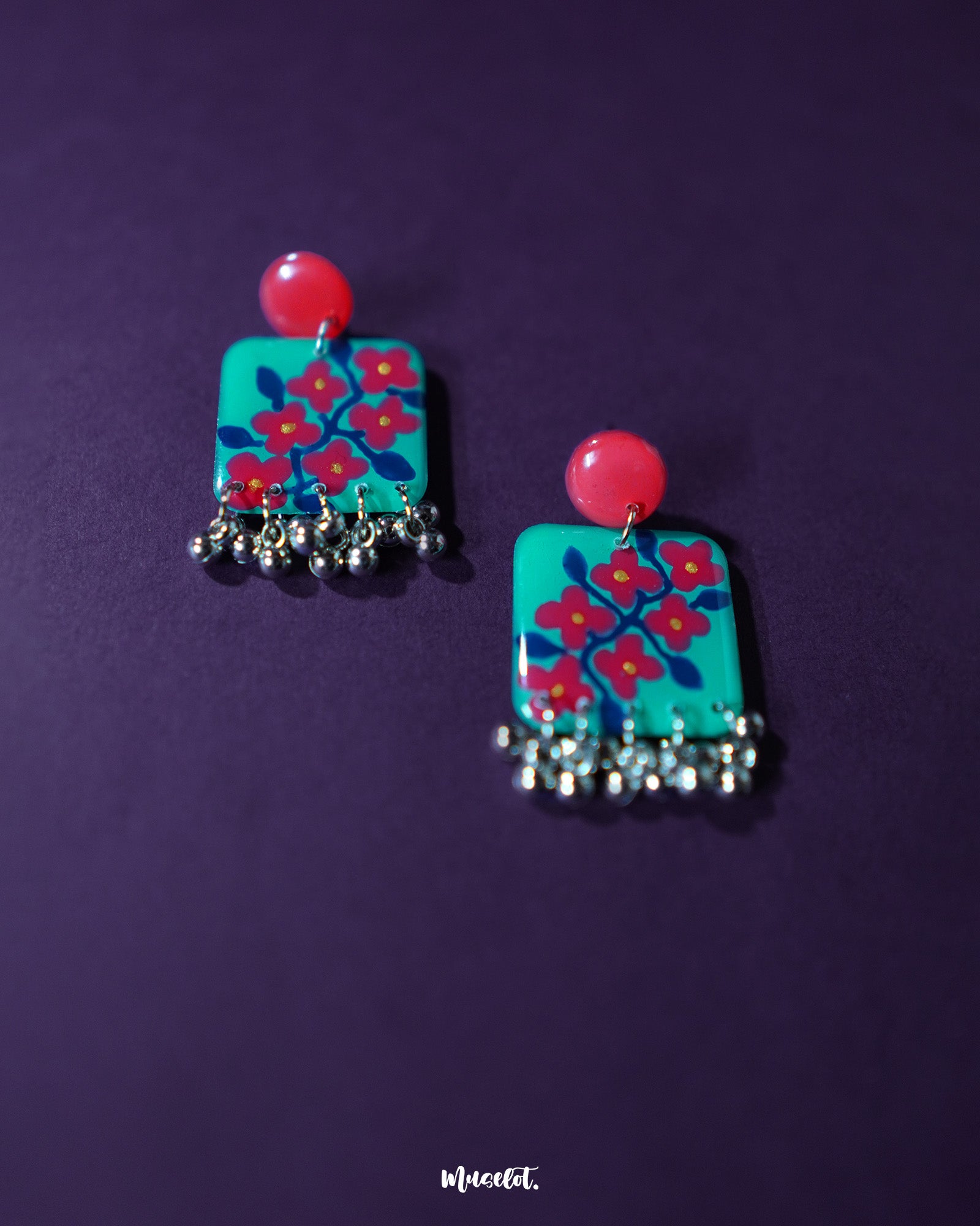 Surkhili floral earrings laid flat on a deep purple background, showcasing their bold turquoise square design with hand-painted red floral motifs and dangling silver ghungroos — Muselot.