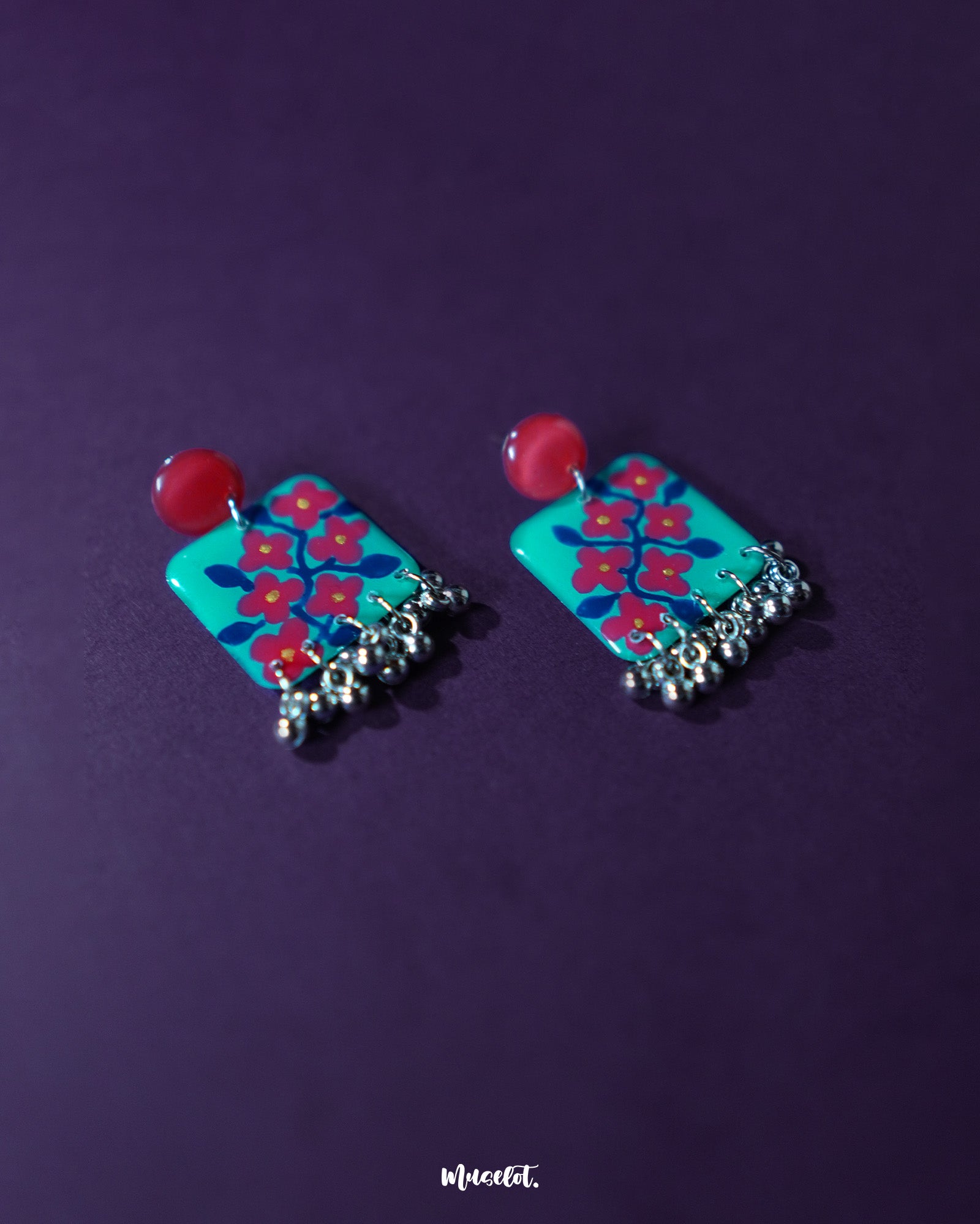 Surkhili floral earrings displayed against a purple background, showcasing the turquoise petals with red floral hand-painting and tiny silver ghungroos hanging below — Muselot.