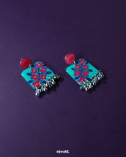 Surkhili floral earrings displayed against a purple background, showcasing the turquoise petals with red floral hand-painting and tiny silver ghungroos hanging below — Muselot.