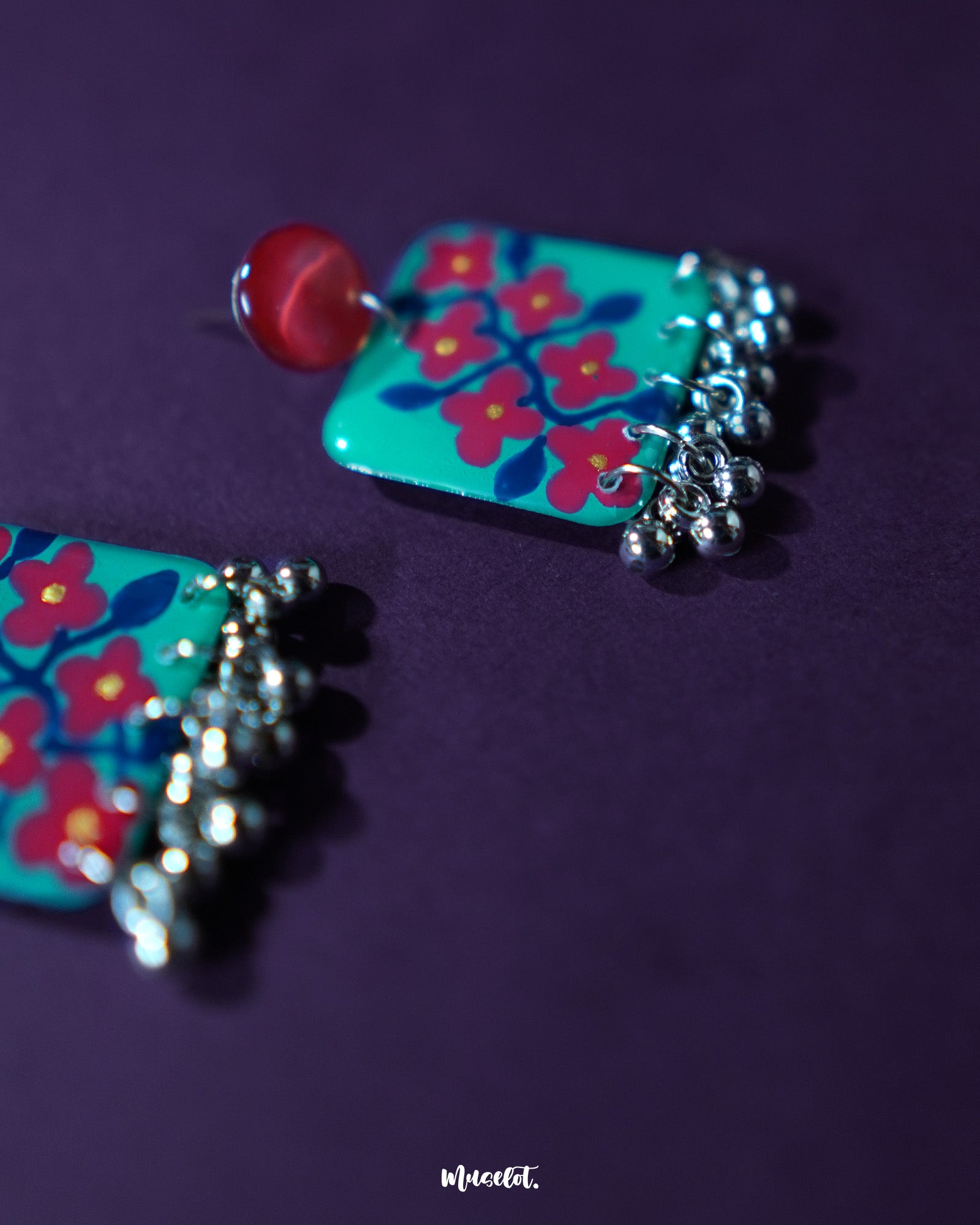 A pair of Surkhili floral earrings laid flat, highlighting the hand-painted red floral details on turquoise petals and delicate silver ghungroos hanging at the bottom — Muselot.