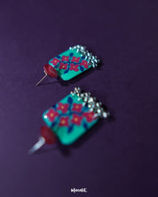 Close-up of Surkhili floral earrings displayed against a neutral backdrop, focusing on the intricate brushwork of the red flower petals and the polished finish of the silver ghungroos — Muselot.
