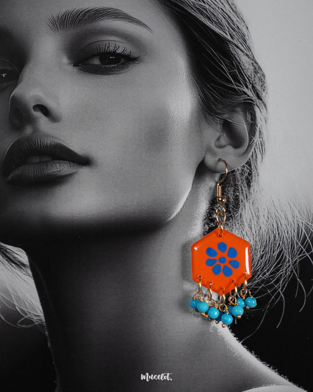 Close-up shot of a model wearing Surkhshaam orange floral hook earrings, featuring vibrant blue floral accents and fine detailing that bring out the bold and elegant charm of the design — Muselot.