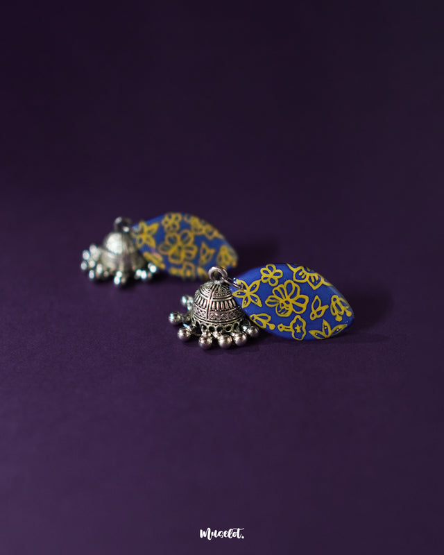 The Tabeer blue jhumkas set against a dark backdrop bring a refreshing pop of color with their floral dome design, showcasing intricate patterns that enhance their elegance — Muselot.