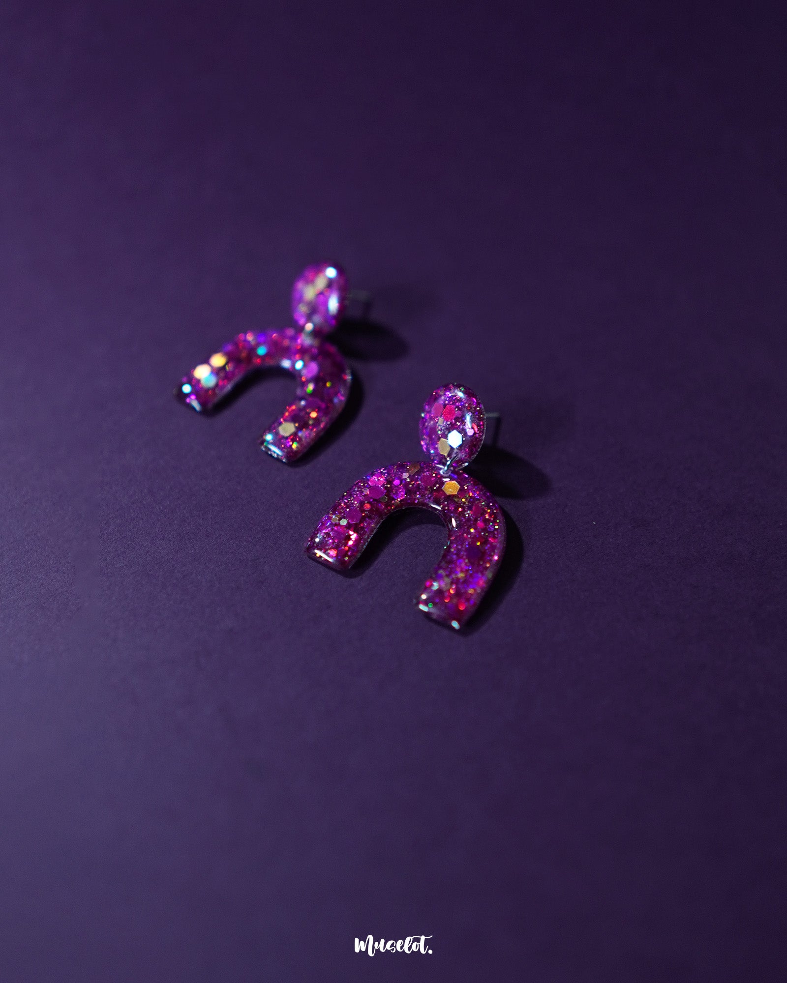 Ulfat glitter stud earrings in vibrant pink and purple resin, displayed against a purple background to showcase their bold arch shape and sparkling finish – Muselot.