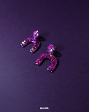 Ulfat glitter stud earrings in vibrant pink and purple resin, displayed against a purple background to showcase their bold arch shape and sparkling finish – Muselot.