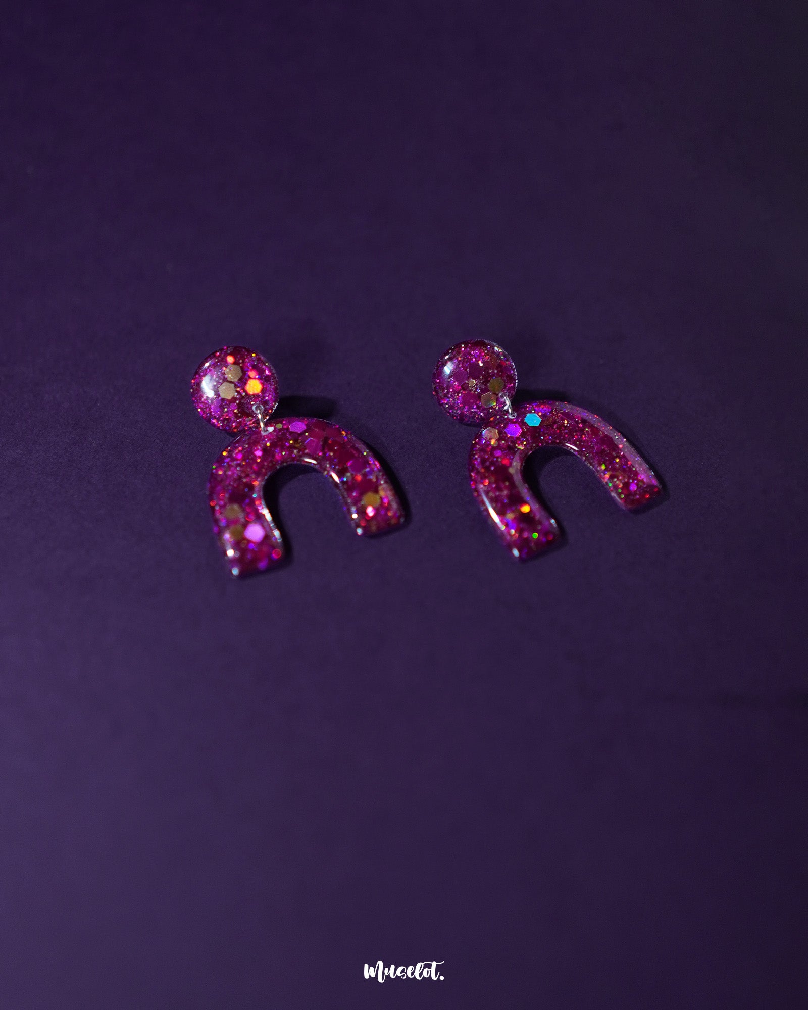 Angled view of Ulfat glitter stud earrings, highlighting the chunky pink-purple sparkles and glossy resin arch design against a plain background – Muselot.