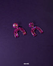 Angled view of Ulfat glitter stud earrings, highlighting the chunky pink-purple sparkles and glossy resin arch design against a plain background – Muselot.