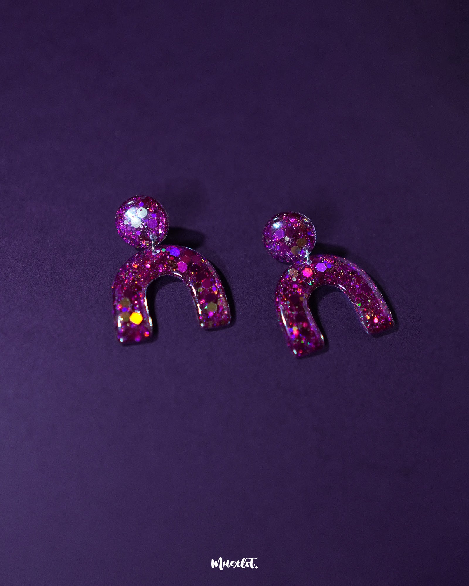 Pair of Ulfat glitter stud earrings laid flat, displaying the vivid pink-purple sparkles and arched resin design on a neutral backdrop – Muselot.