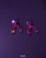 Pair of Ulfat glitter stud earrings laid flat, displaying the vivid pink-purple sparkles and arched resin design on a neutral backdrop – Muselot.