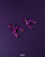 Detailed close-up of Ulfat glitter stud earrings, highlighting the embedded pink and purple sparkles within the glossy resin arch shape – Muselot.