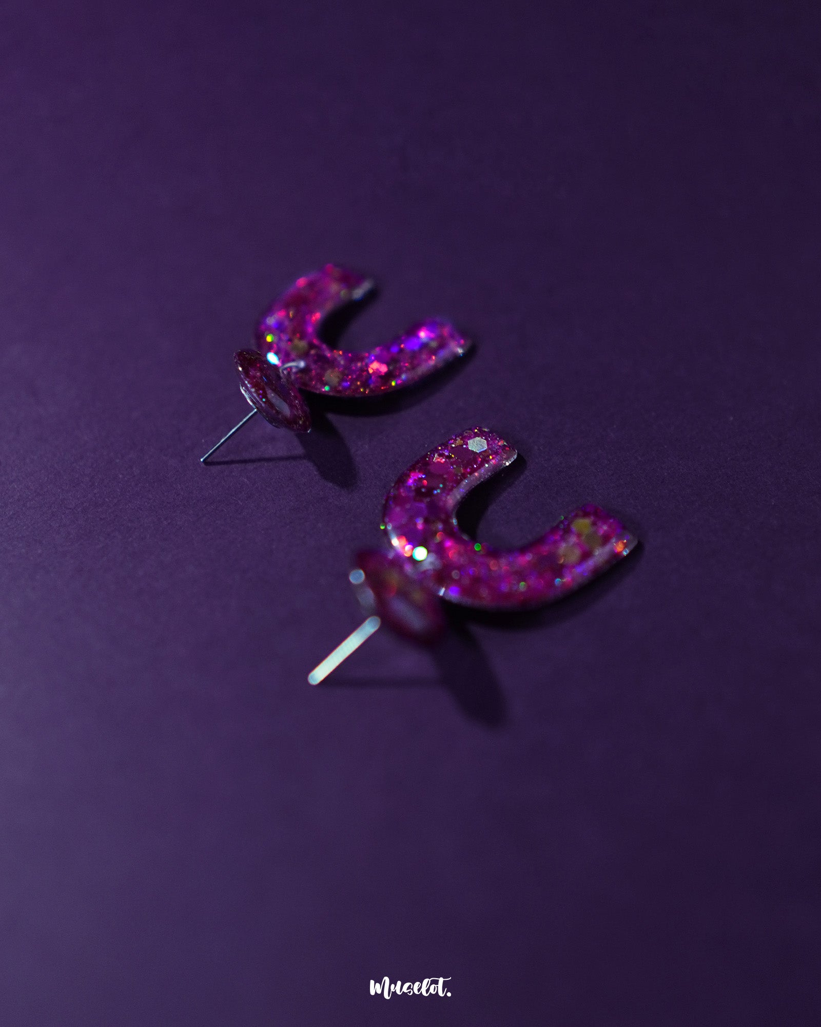 Ulfat glitter stud earrings displayed upright, showcasing the arch silhouette and vibrant pink-purple glitter blend against a simple backdrop – Muselot.