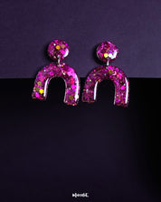 Ulfat glitter resin stud earrings in vibrant pink-purple hues, featuring a bold arch shape adorned with chunky sparkles, displayed on a dark background – Muselot.