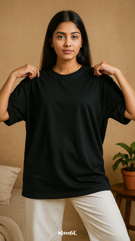 A model wearing a unisex oversized black T-shirt gently adjusting the collar, showcasing the minimal lifestyle appeal and effortless charm of this versatile wardrobe essential tee for an everyday wear — Muselot.