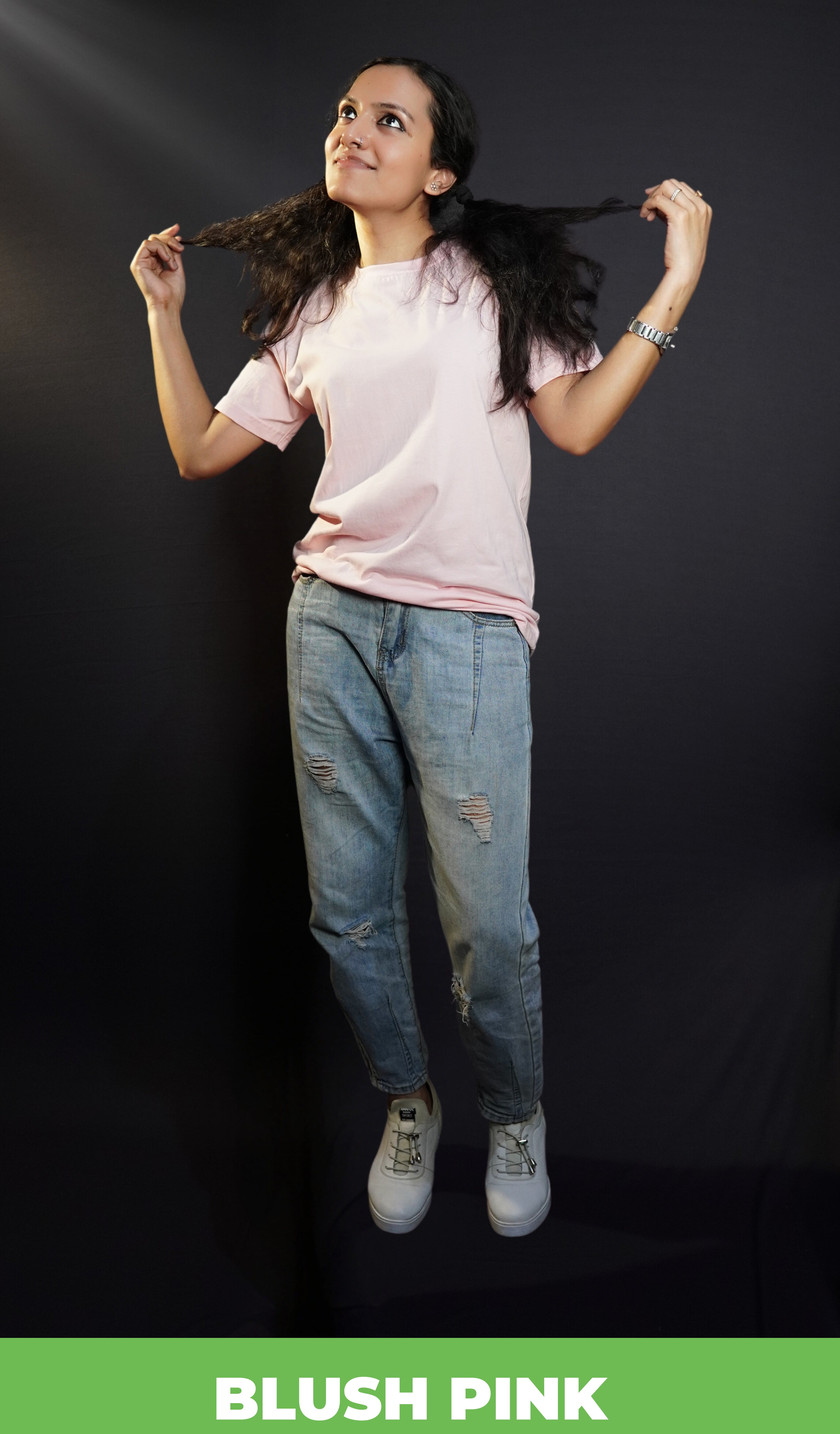 A stylish short-haired model poses confidently in a round neck plain blush pink t shirt and ripped blue jeans against a dark background, pulling the tips of her hair at the sides with both hands, showcasing a trendy snug fit-muselot