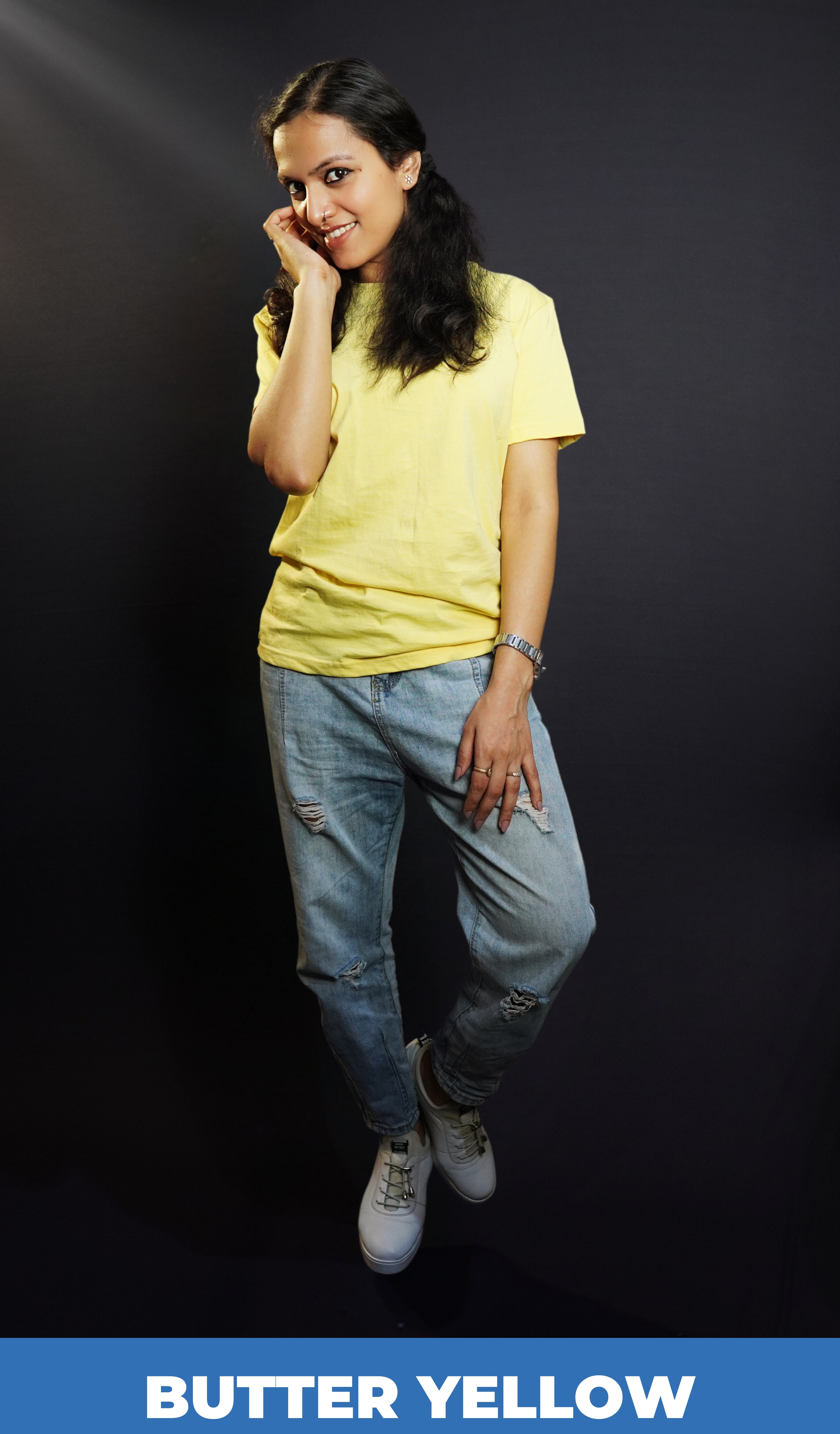 A woman posing happily as she stands against a dark background with her hand on the side of her face and another on her thigh, wearing a butter yellow plain long length stretchable t-shirt, showcasing a voguish popular style-Muselot