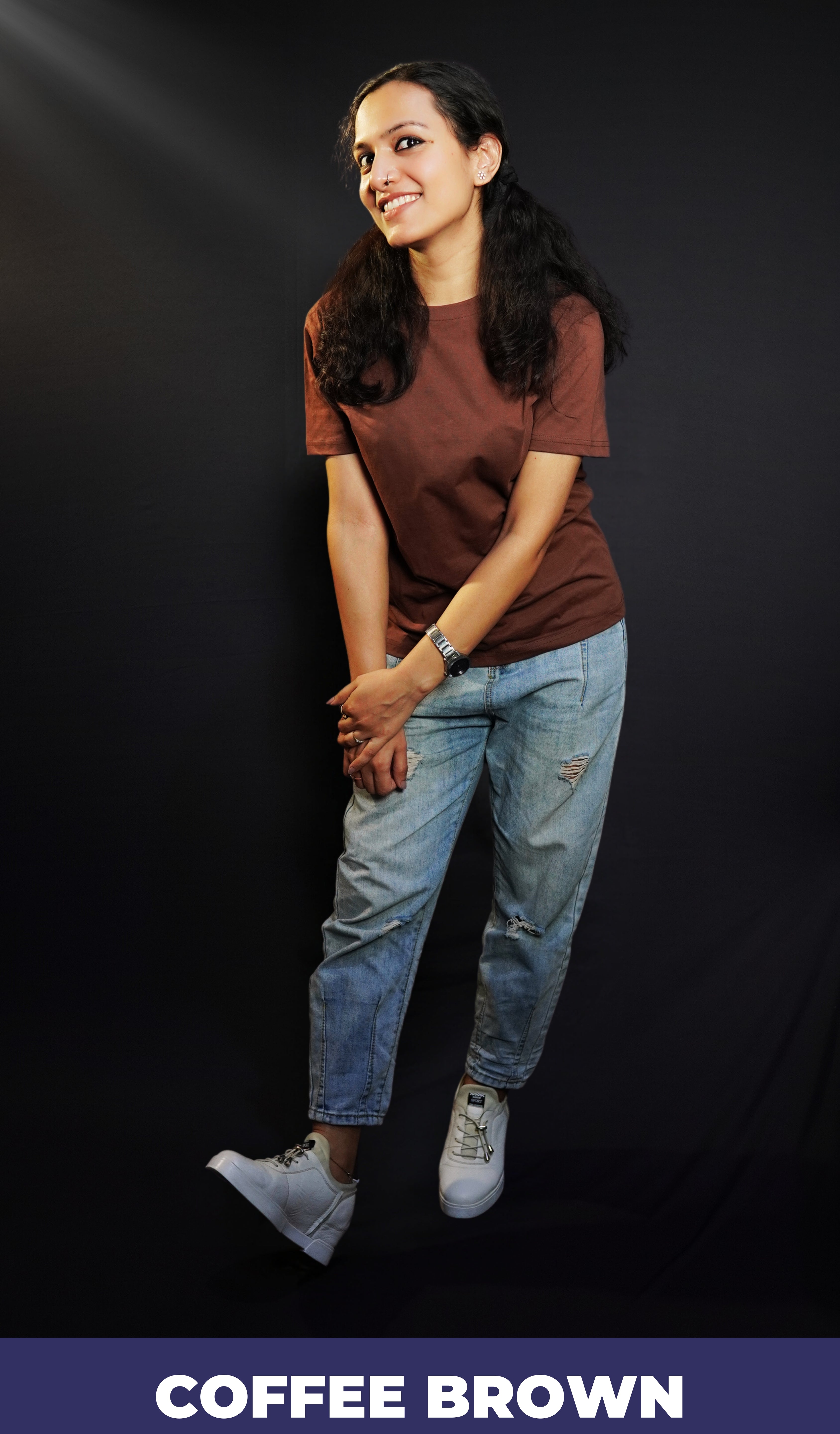A woman in a long length plain coffee brown t-shirt, posing happily as she slightly bends forward with her hands grabbing each other, highlighting a popular cozy fit-Muselot