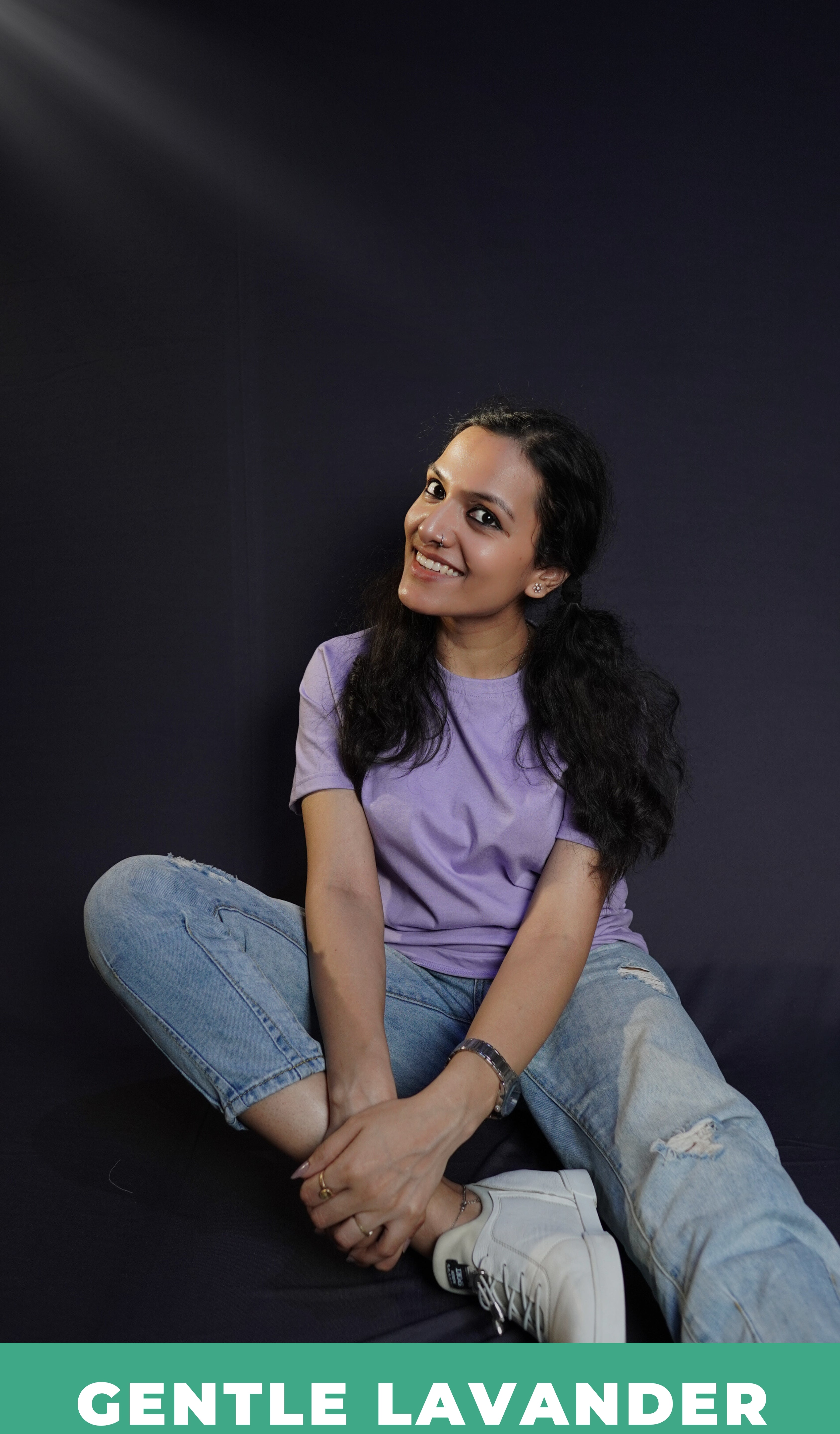 A stylish model wearing a plain short sleeved round neck t-shirt, posing by sitting against a dark background grabbing her leg gently with both hands, showcasing a swank perfect fit-Muselot
