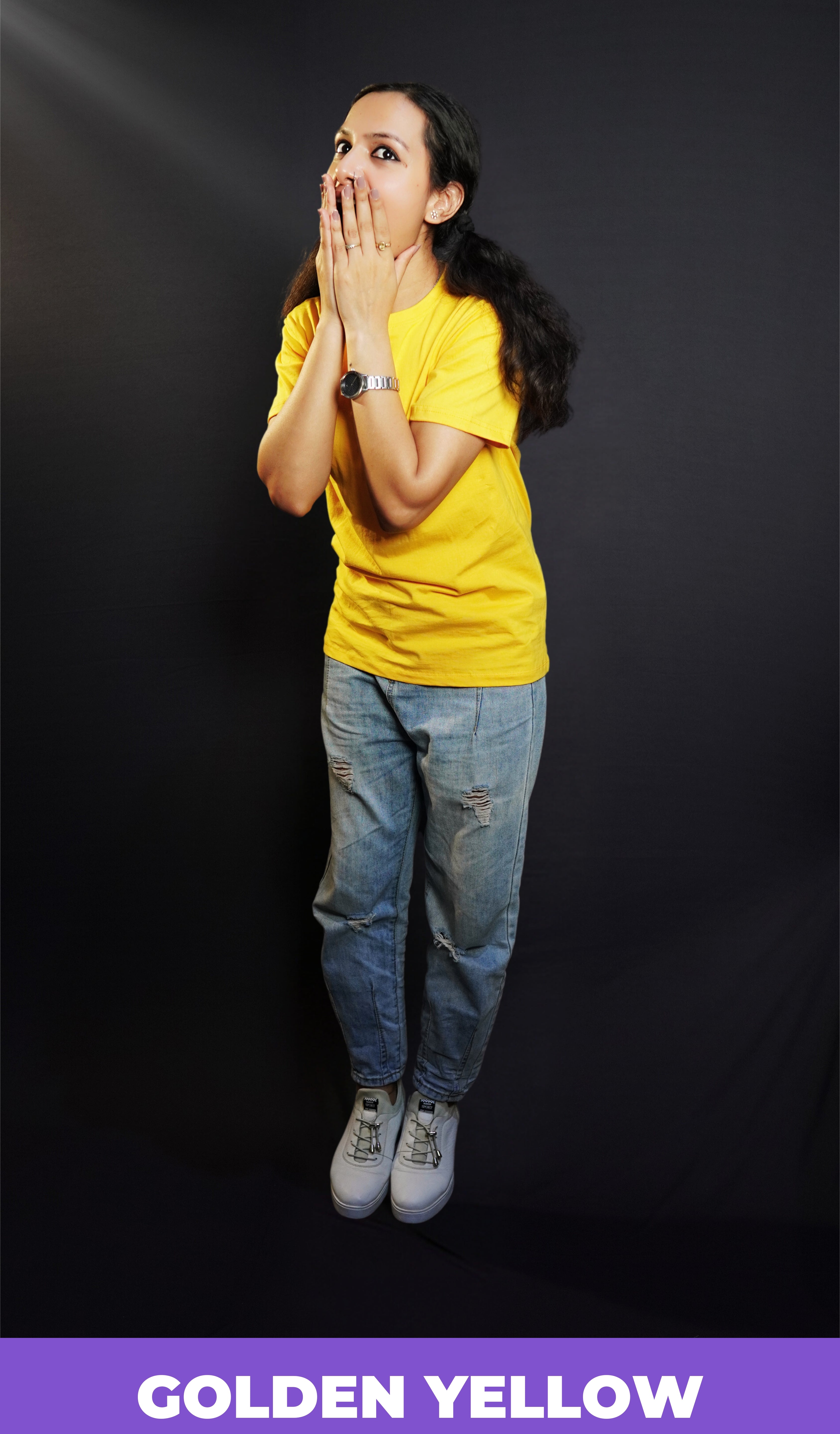 A woman wearing a unisex golden yellow long length T shirt pairing with a blue jeans and sports shoes standing excitedly against a dark background-Muselot
