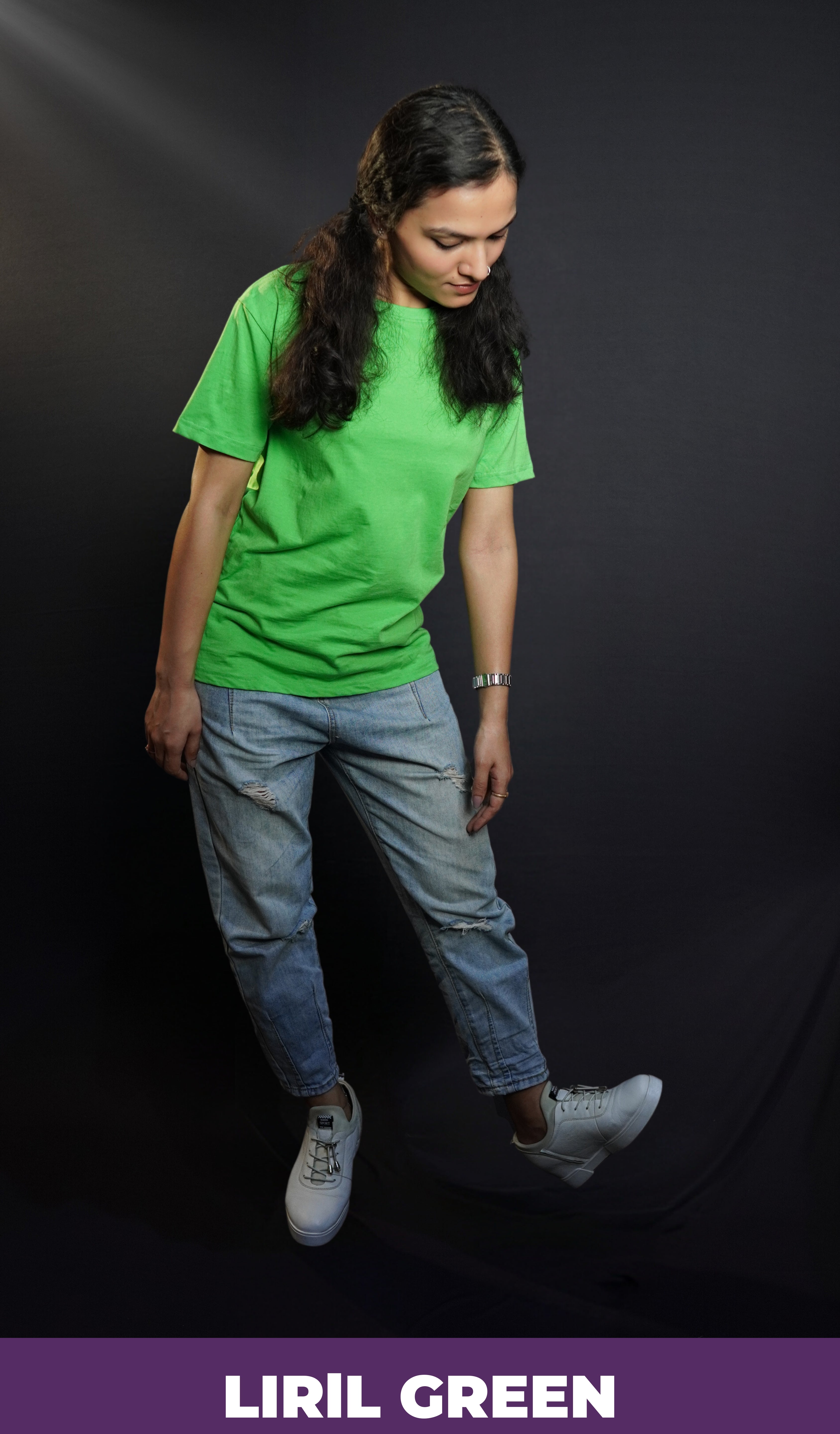 A woman wearing a women's long length short sleeved stretchable liril green t-shirt, posing against a dark background as she bends down slightly, looking at her white shoes, showcasing a relaxed cool fit-Muselot