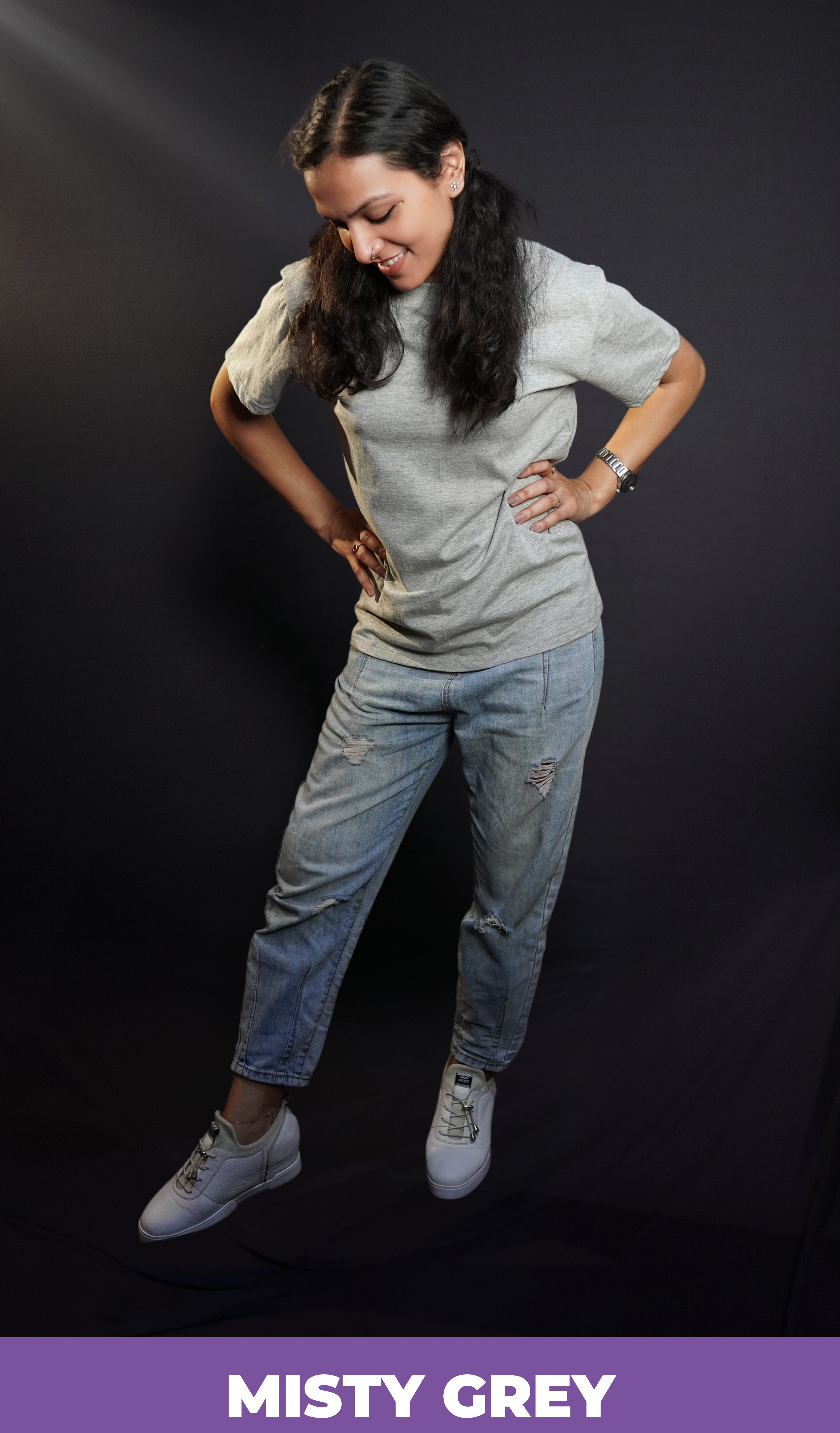 A woman posing with her hands on her waist standing slghtly sideways bending down a little, wearing a plain unisex short sleeved stretchable misty grey t-shirt, highlighting a stylish comfortable fit-Muselot