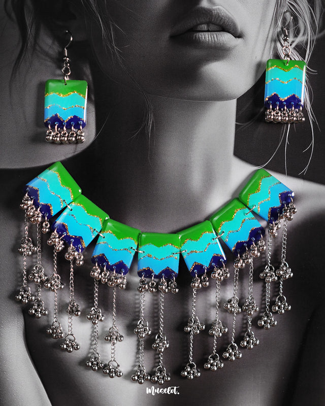 A woman wearing the Waterfall Boho Jewelry Set, with the vibrant enamel necklace resting along her neckline. The bold green, turquoise, and navy segments with glittery zigzag accents, paired with cascading silver-toned chains and beads, create a striking contrast against her outfit, showcasing the statement design's artistic and bohemian appeal — Muselot.