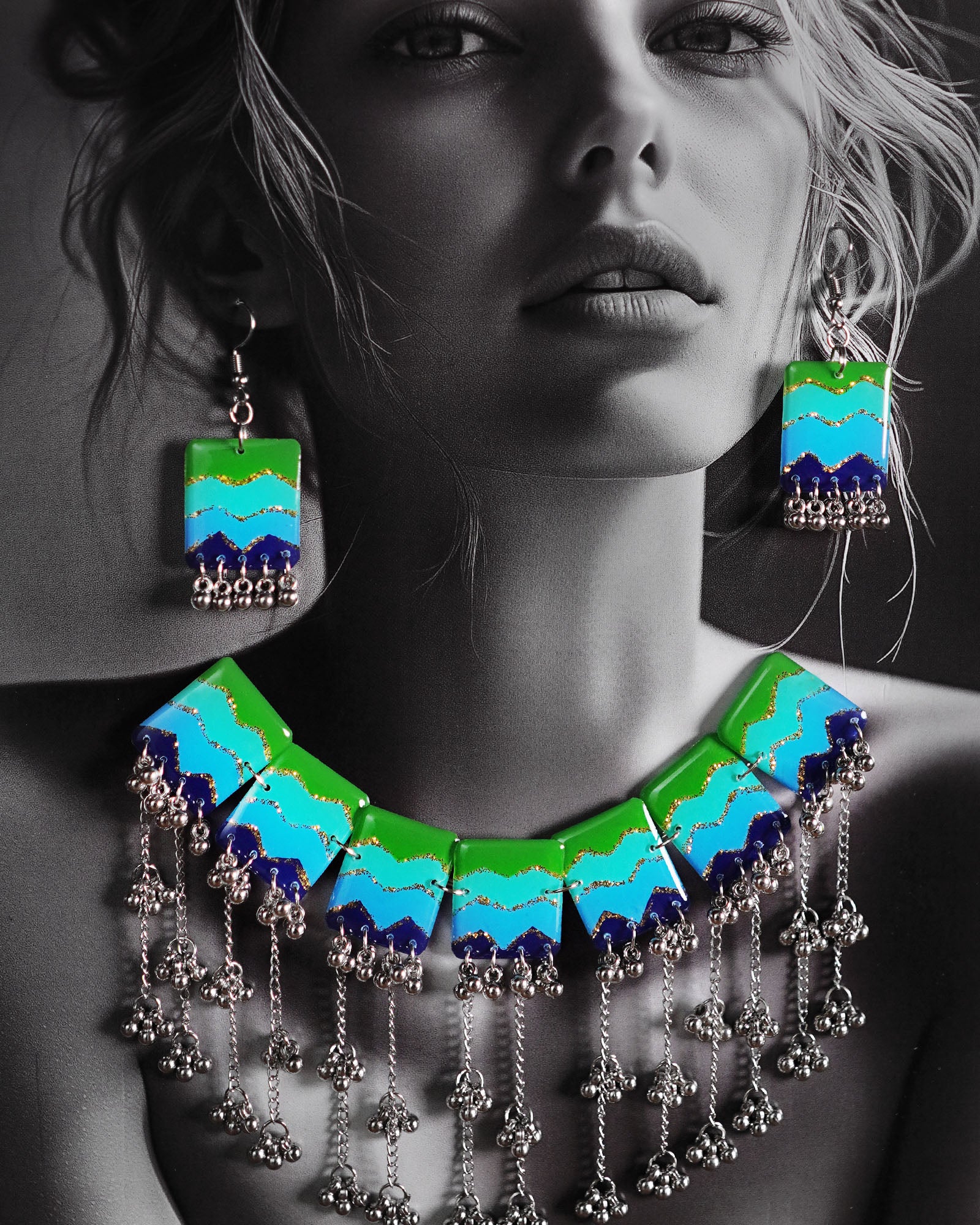 A model adorned in the Boho Jewelry Set, featuring a bold necklace with layered green, aqua, and deep blue panels highlighted with glitter accents. Silver-toned dangling chains and clustered beads sway below the enamel plates, adding movement and charm to the piece — Muselot.