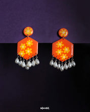 Zafraan floral earrings featuring round orange studs with yellow flower motifs, attached to hexagonal orange drops adorned with matching floral designs and finished with dangling white pearl-like beads, beautifully displayed against a deep purple background – Muselot.