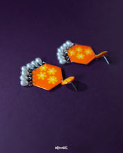 Zafraan floral earrings displayed on a purple background, featuring round orange studs with yellow flower motifs attached to hexagonal orange drops adorned with matching floral designs, finished with dangling white pearl-like beads – Muselot.