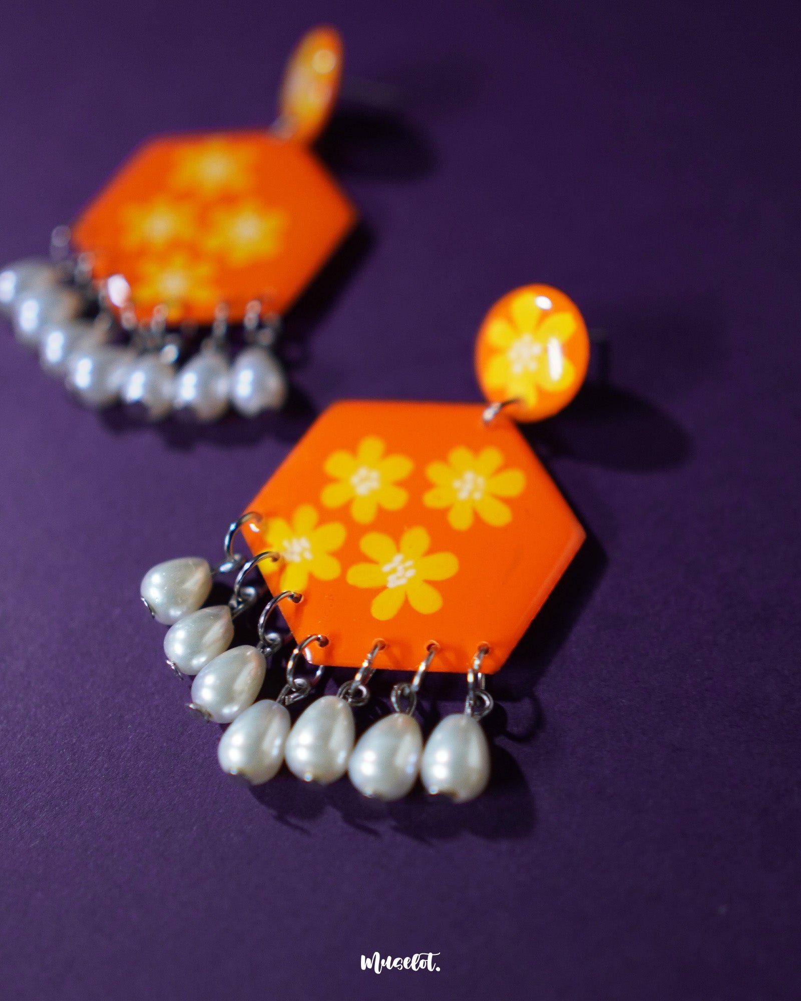 Close-up view of the Zafraan floral earrings, featuring round orange studs with yellow flower motifs attached to hexagonal orange drops with matching floral patterns, accented by dangling white pearl-like beads, capturing their vivid colors and fine craftsmanship – Muselot.