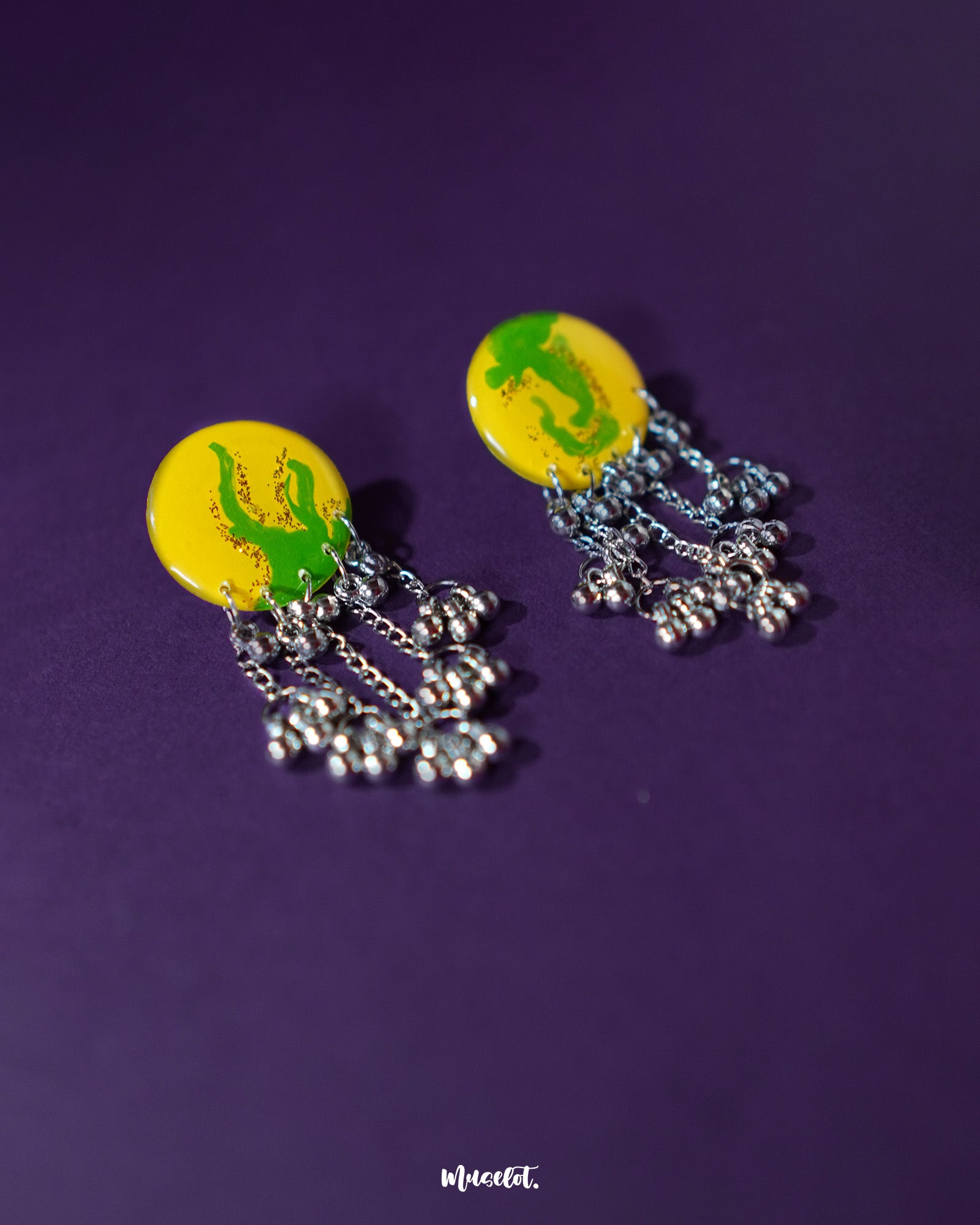 Zarangi yellow drop earrings displayed against a deep purple background, featuring round yellow tops with green accents and cascading silver chains adorned with small metallic ghungroos — Muselot.