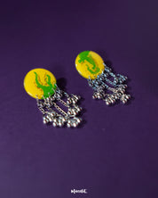 Zarangi yellow drop earrings displayed against a deep purple background, featuring round yellow tops with green accents and cascading silver chains adorned with small metallic ghungroos — Muselot.