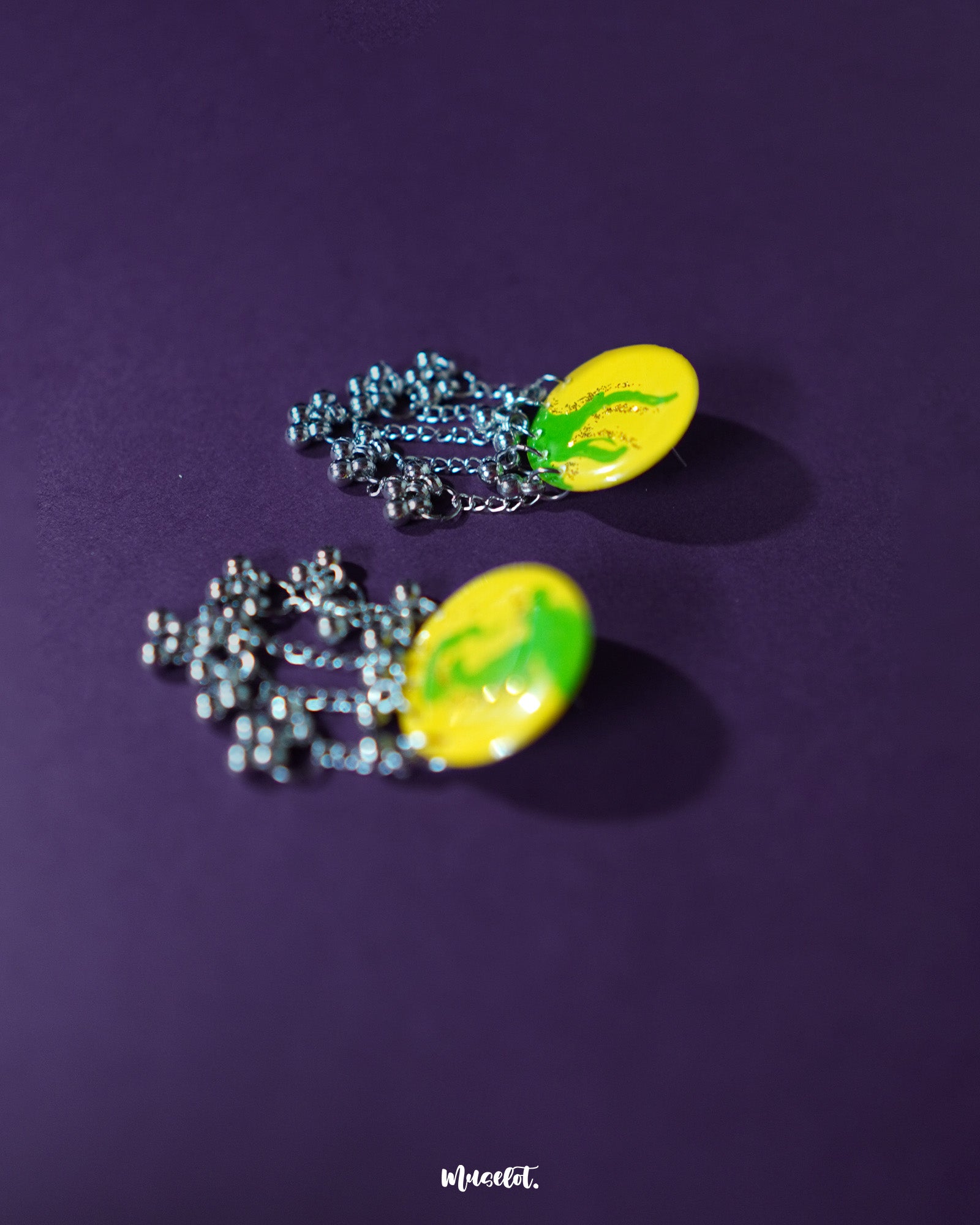 Zarangi yellow drop earrings displayed against a purple background, showcasing their vibrant circular tops with green floral accents and dangling silver chains adorned with metallic beads — Muselot.