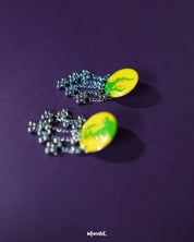 Zarangi yellow drop earrings displayed against a purple background, showcasing their vibrant circular tops with green floral accents and dangling silver chains adorned with metallic beads — Muselot.