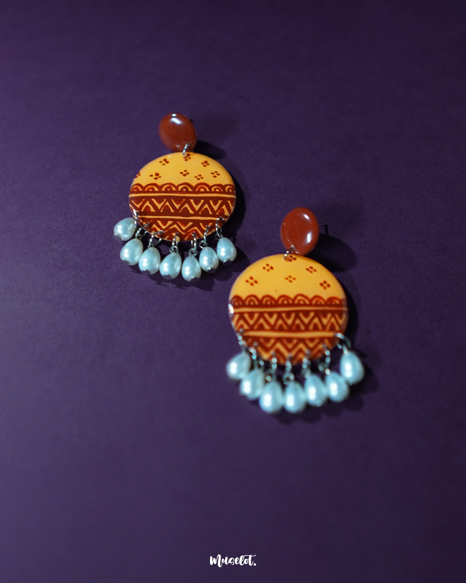 Zardozi traditional handmade earrings against a purple background, showcasing mustard yellow discs adorned with rust-hued hand-painted ethnic patterns, finished with dangling white pearl drops—highlighting the elegance of handcrafted heritage — Muselot.