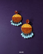 Zardozi traditional handmade earrings against a purple background, showcasing mustard yellow discs adorned with rust-hued hand-painted ethnic patterns, finished with dangling white pearl drops—highlighting the elegance of handcrafted heritage — Muselot.