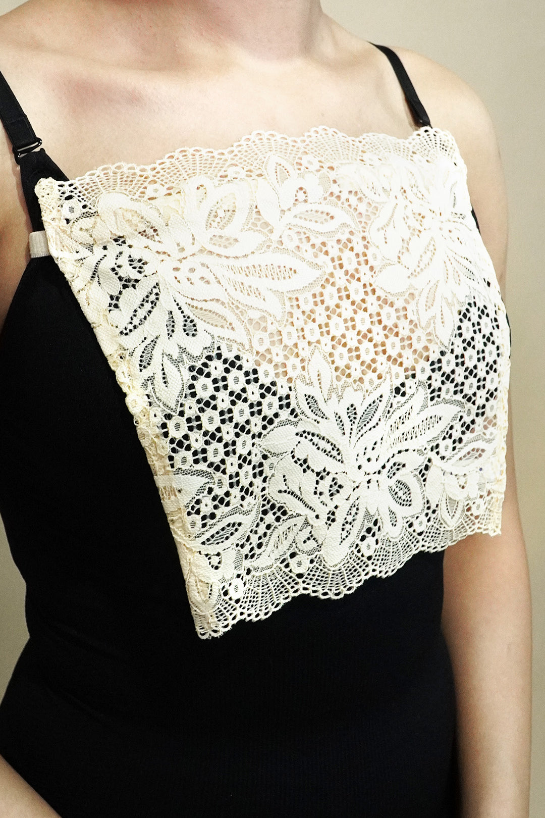side close up of woman wearing beige lace camisole insert over black top on beige background, highlighting broad lace panel designed for better coverage on deep neck outfits – muselot