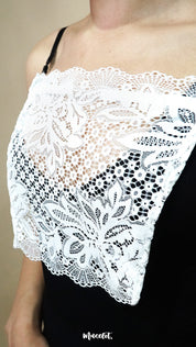 Side-angled close up of white lace camisole insert worn over black dress on beige background, showing broad lace design created to hide low necklines smoothly – muselot