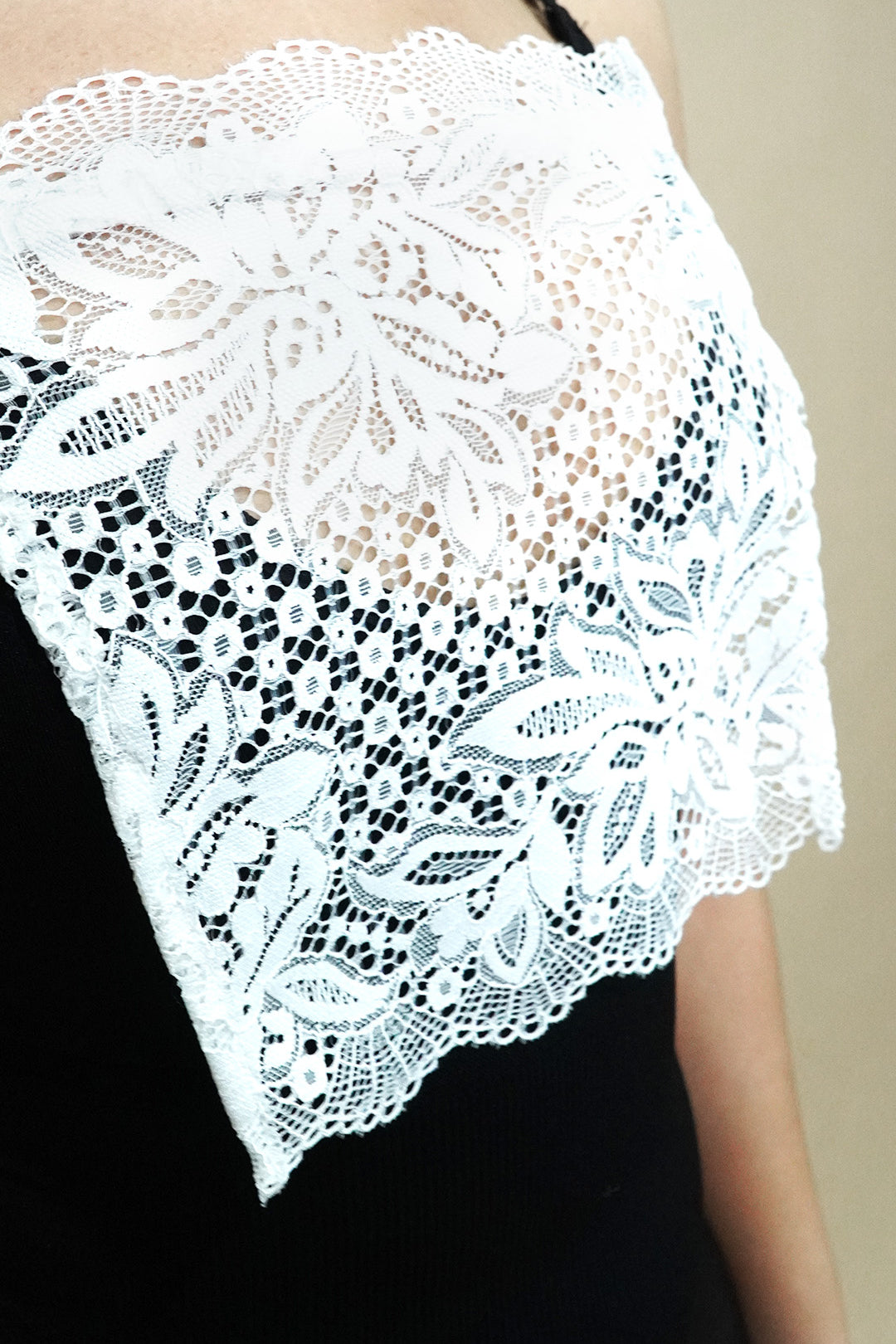 Side-angled close up of white lace camisole insert worn over black dress on beige background, showing broad lace design created to hide low necklines smoothly – muselot