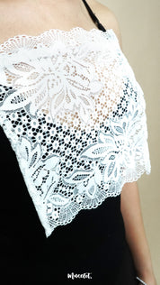 Side-angled close up of white lace camisole insert worn over black dress on beige background, showing broad lace design created to hide low necklines smoothly – muselot
