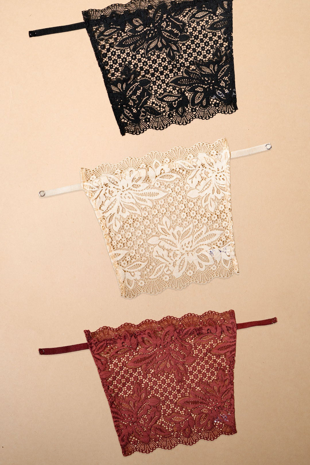 Three lace clip-on neckline covers in black, beige and burgundy displayed on a beige background, featuring rich floral lace patterns, adjustable straps and elegant neckline coverage. Perfect for deep neck tops, dresses and blouses, these breathable mock camisole inserts add a sophisticated layered look while providing comfortable coverage without adding bulk – Muselot