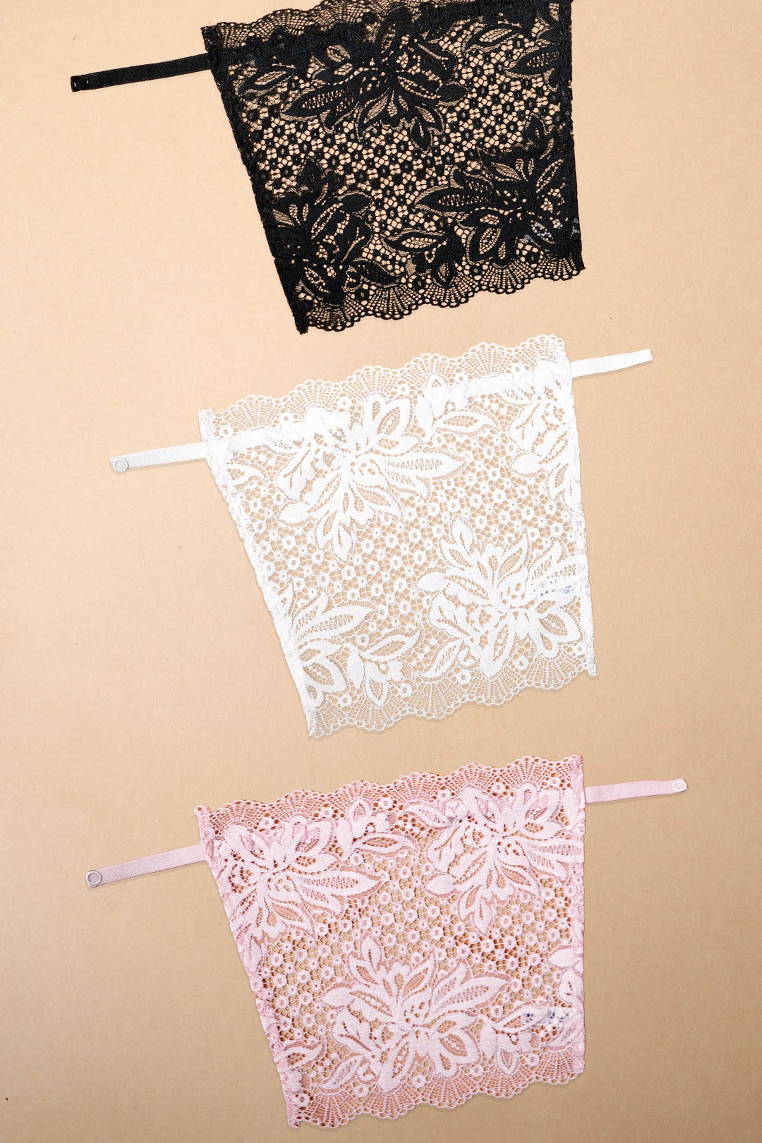 A pack of three clip-on neckline covers in black, white and baby pink is displayed on a beige background. Crafted with premium floral lace and adjustable straps, these deep neck cover-ups add elegant neckline coverage to dresses, tops, kurtis and blouses. The lightweight lace design blends seamlessly with different necklines, offering a stylish modesty panel, layering accessory and feminine fashion accent without the need for a full camisole – Muselot