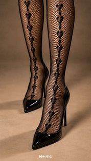 A close-up shot of a woman wearing black fishnet garter stockings with glossy pointed heels, highlighting the repeating heart motif, fine fishnet texture and detailed pattern placement along the lower legs - Muselot