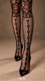 A close-up shot of a woman wearing black fishnet garter stockings with glossy black pointed heels, highlighting the vertical heart motif, floral lace detailing and fine mesh texture along the legs - Muselot