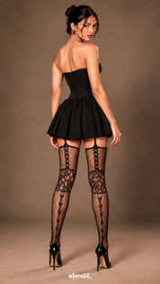 A woman shown from the back wearing black fishnet garter stockings with a black mini dress and heels, showcasing the rear view of the garter straps, floral lace pattern and vertical heart detailing - Muselot